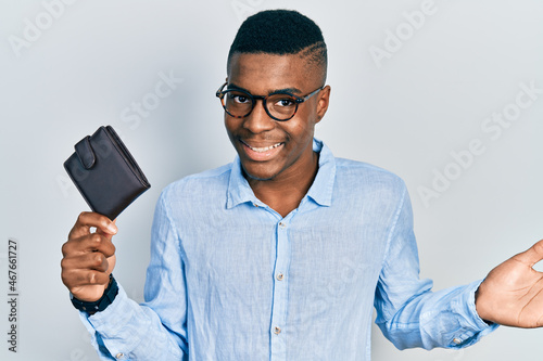 Young african american man wearing glasses holding leather wallet celebrating achievement with happy smile and winner expression with raised hand © Krakenimages.com