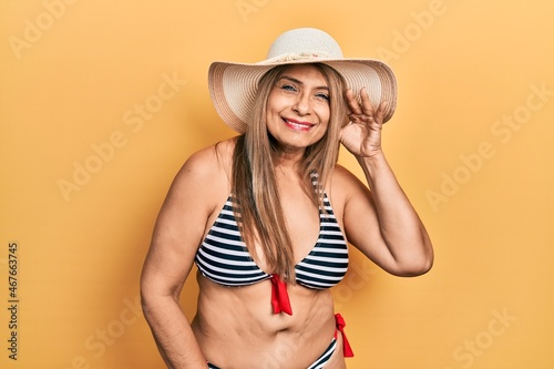Middle age hispanic woman wearing bikini and summer hat smiling with hand over ear listening an hearing to rumor or gossip. deafness concept.