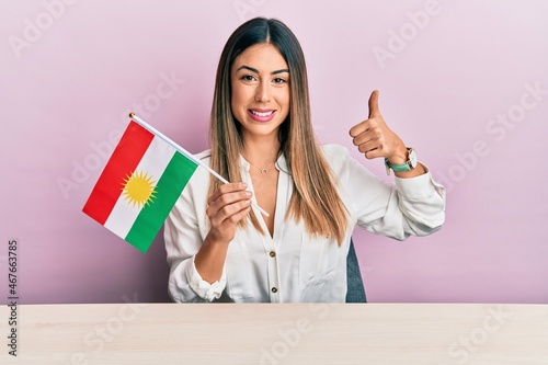 Young hispanic woman holding kurdistan flag sitting on the table smiling happy and positive, thumb up doing excellent and approval sign photo