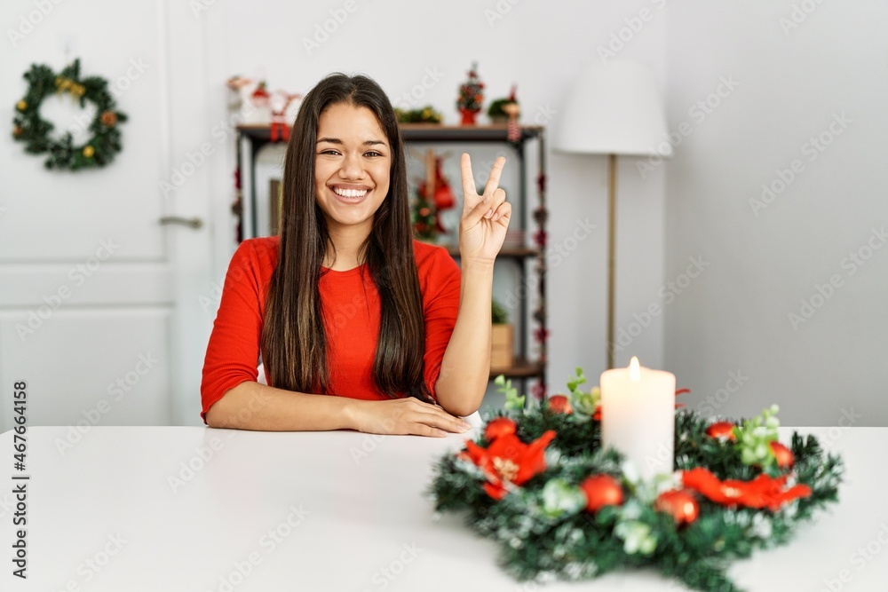Young brunette woman sitting on the table on christmas smiling with happy face winking at the camera doing victory sign. number two. Young brunette woman sitting on the table on christmas smiling with happy face winking at the camera doing victory sign. number two.