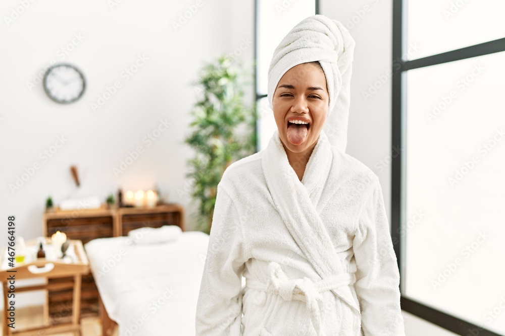 Young brunette woman wearing towel and bathrobe standing at beauty center sticking tongue out happy with funny expression. emotion concept. Young brunette woman wearing towel and bathrobe standing at beauty center sticking tongue out happy with funny expression. emotion concept.