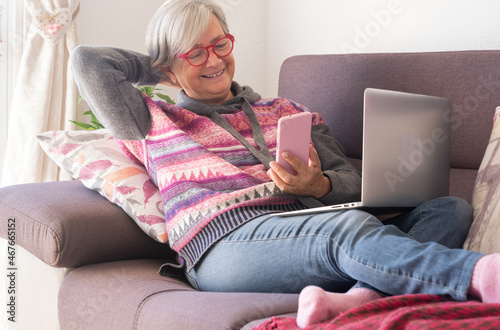 Attractive senior woman resting on sofa at home using laptop and mobile phone. Smiling elderly retiree enjoying tech and social