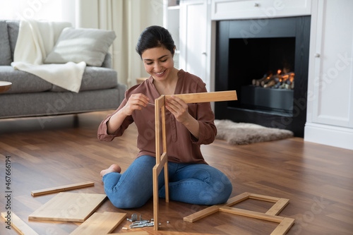 Woman assembling new furniture at home, new purchase, hobby concept. Young Indian female sit on warm floor in living room with fireplace holding wooden boards chair details, collects modern furniture