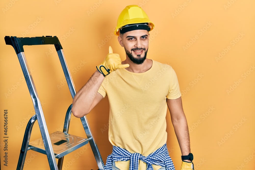 Handsome man with beard by construction stairs wearing hardhat smiling doing phone gesture with hand and fingers like talking on the telephone. communicating concepts. Handsome man with beard by construction stairs wearing hardhat smiling doing phone gesture with hand and fingers like talking on the telephone. communicating concepts.