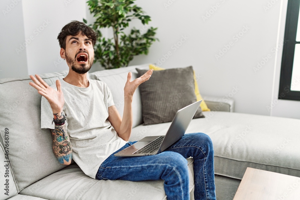 Hispanic man with beard sitting on the sofa crazy and mad shouting and yelling with aggressive expression and arms raised. frustration concept.