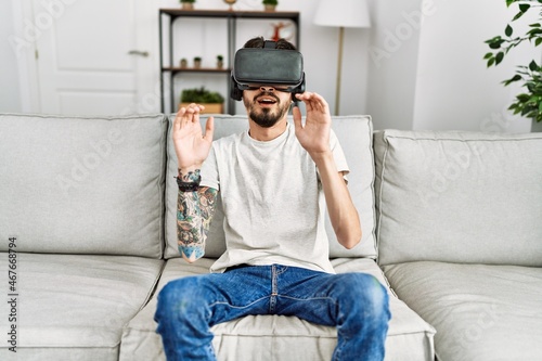 Young hispanic man playing video game using virtual reality glasses at home.