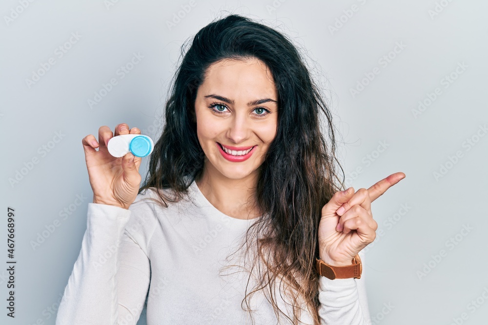 Young hispanic woman holding contact lenses smiling happy pointing with hand and finger to the side Young hispanic woman holding contact lenses smiling happy pointing with hand and finger to the side