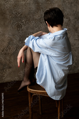 Young woman in stockings and blue oversize shirt sits on chair legs tucked up and facing the wall
