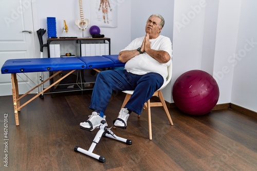 Senior caucasian man at physiotherapy clinic using pedal exerciser begging and praying with hands together with hope expression on face very emotional and worried. begging.