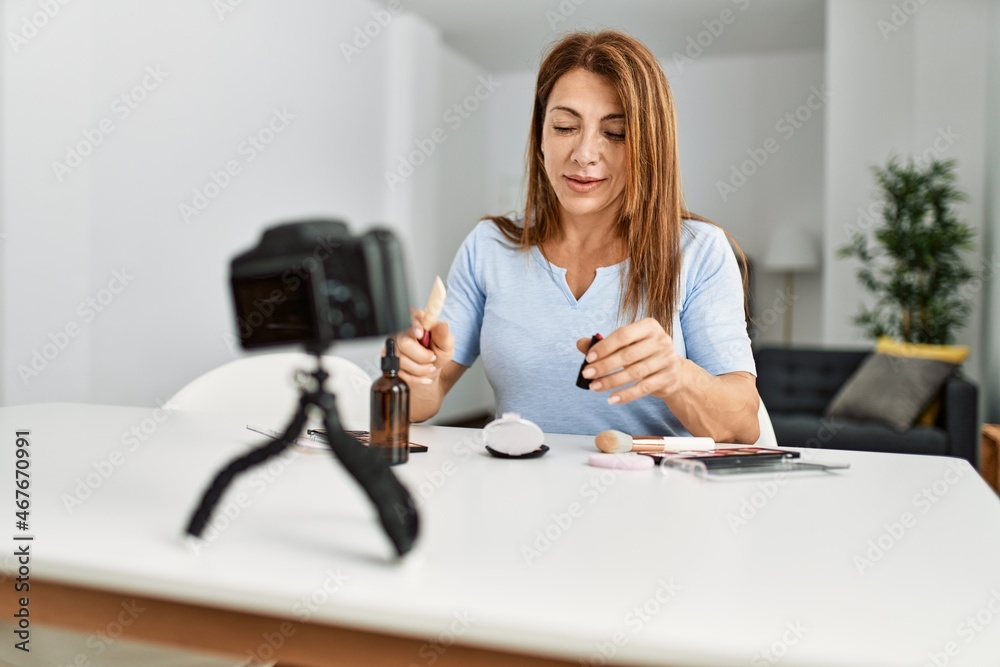 Middle age caucasian woman making makeup tutorial using camera sitting on the table at home. Middle age caucasian woman making makeup tutorial using camera sitting on the table at home.