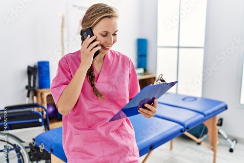 Young caucasian woman wearing physiotherapist uniform talking on the smartphone at physiotherapy clinic photo