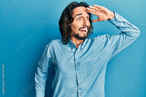 Young hispanic man wearing casual clothes very happy and smiling looking far away with hand over head. searching concept. © Krakenimages.com