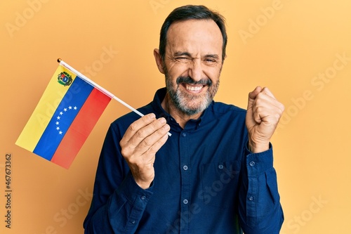 Middle age hispanic man holding venezuelan flag screaming proud, celebrating victory and success very excited with raised arm