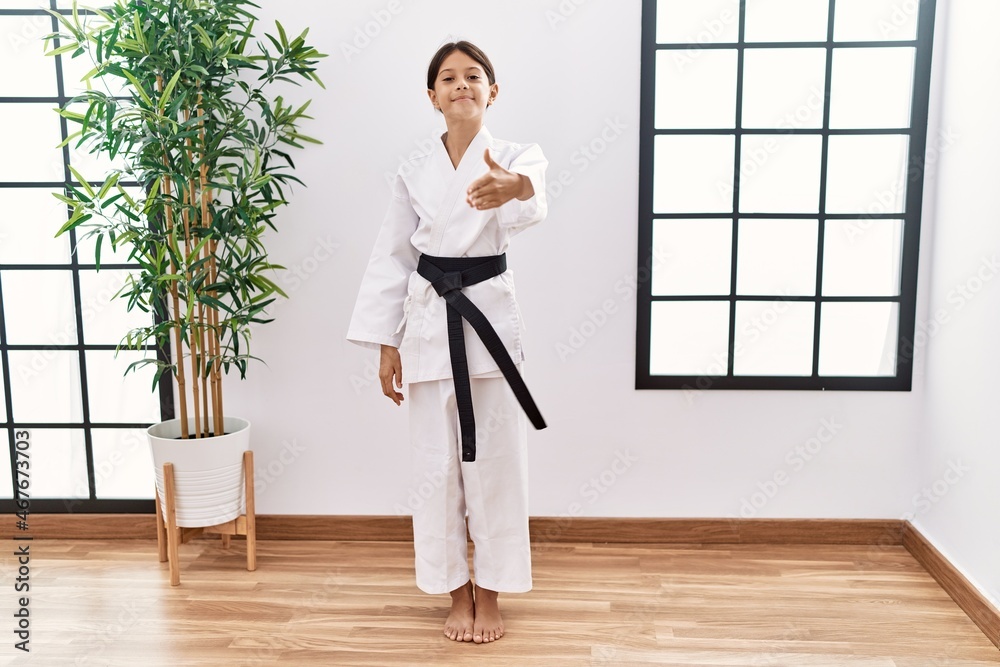 Young hispanic girl wearing karate kimono and black belt smiling friendly offering handshake as greeting and welcoming. successful business.