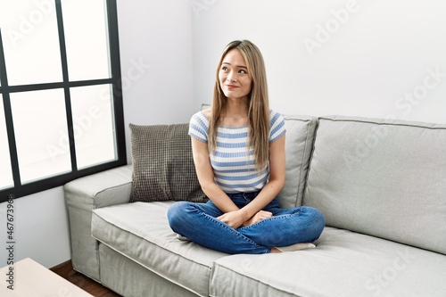 Asian young woman sitting on the sofa at home smiling looking to the side and staring away thinking.