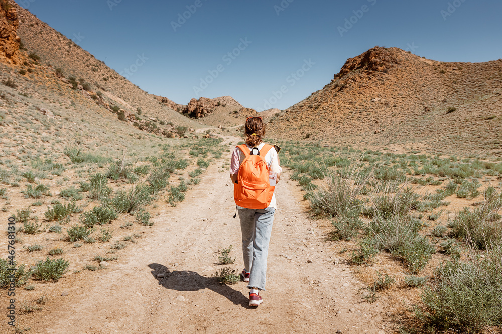 A woman with a backpack walks along a trail in a deserted canyon with red rocks. Hiking path and the dangers of solo trekking