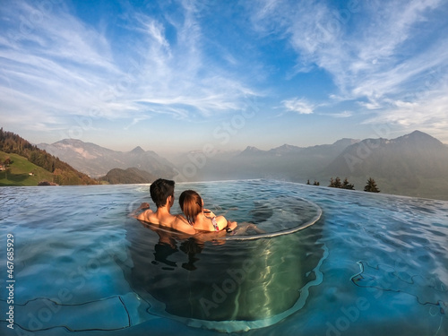 Back of young romantic Asian couple sitting together on an underwater seat in the swimming pool admired beautiful seascapes surrounded by mountains with amazing blue sky and soft clouds on a clear day
