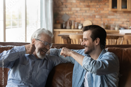Give me five dad. Laughing old father bumping fists with adult son sitting on sofa at home having fun talking joking relaxing on weekend. Happy aged dad support grown kid encourage greet with success