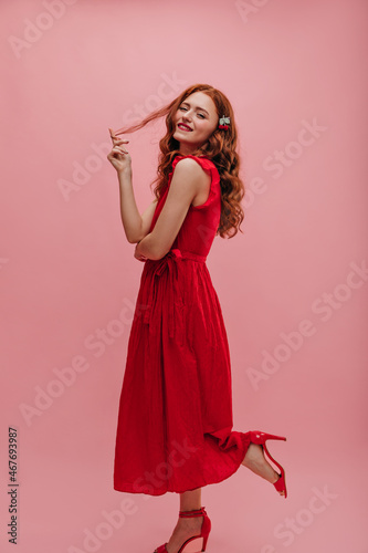 Full length photo seductive caucasian woman in red long dress on pink background. Beauty with arms crossed winds red hair on finger biting lip with red lipstick. Lifestyle leisure and beauty concept.