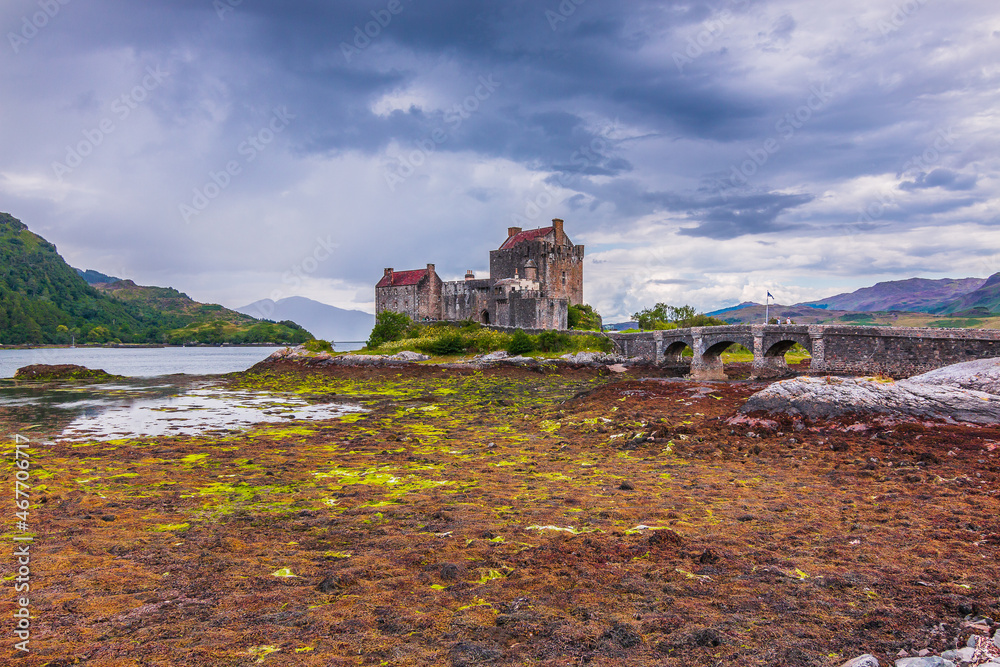 Lake Loch Duich at low tide. Eilean Donan Castle in summer on the island. Castle and a stone bridge and small archways. Algae on stones in the foreground and hills in the background with clouds