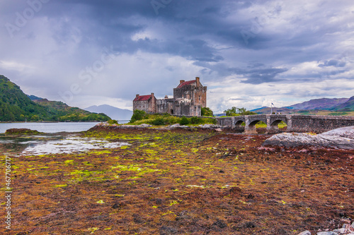 Lake Loch Duich at low tide. Eilean Donan Castle in summer on the island. Castle and a stone bridge and small archways. Algae on stones in the foreground and hills in the background with clouds