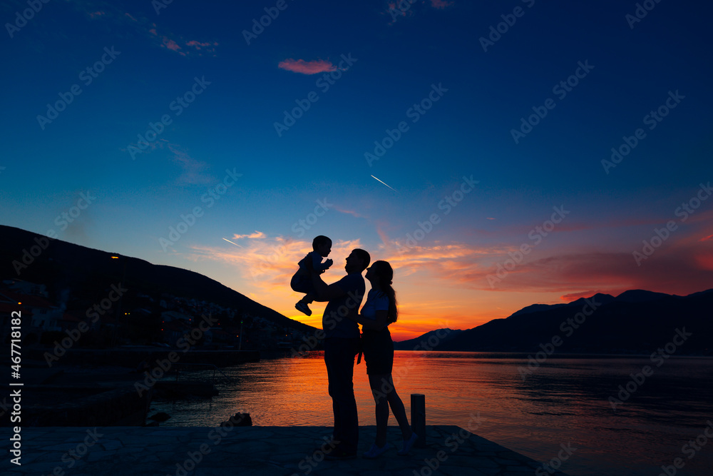 Silhouettes of father holding small child and mother standing next to them on the pier against the backdrop of the sea, mountains and houses at sunset