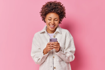 Happy pretty young woman holds mobile phone makes shopping online enjoys best offer at sale from store types text message wears warm fur jacket isolated over pink background surfs social media