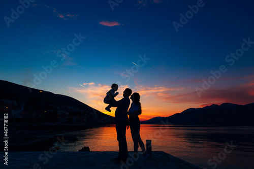 Silhouettes of father holding small child and mother standing next to them on the pier against the backdrop of the sea, mountains and houses at sunset