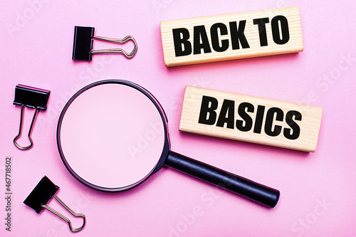 On a pink background, a magnifier, black paper clips and wooden blocks with the text BACK TO BASICS. Business concept