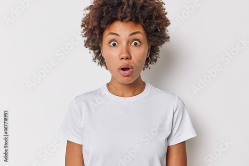 Photo of startled curly haired woman holds breath has eyes popped out cannot believe in shocking news wears casual t shirt isolated over white background stands anxious. Human reactions concept