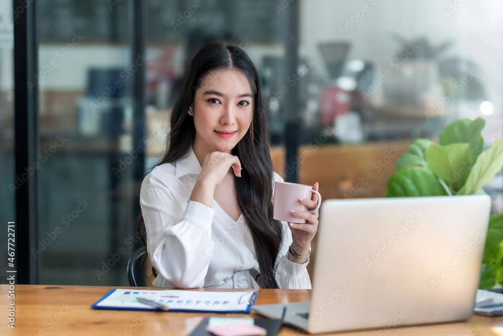 Smiling young Asian businesswoman holding a coffee mug and laptop at the office. Looking at the camera. Smiling young Asian businesswoman holding a coffee mug and laptop at the office. Looking at the camera.