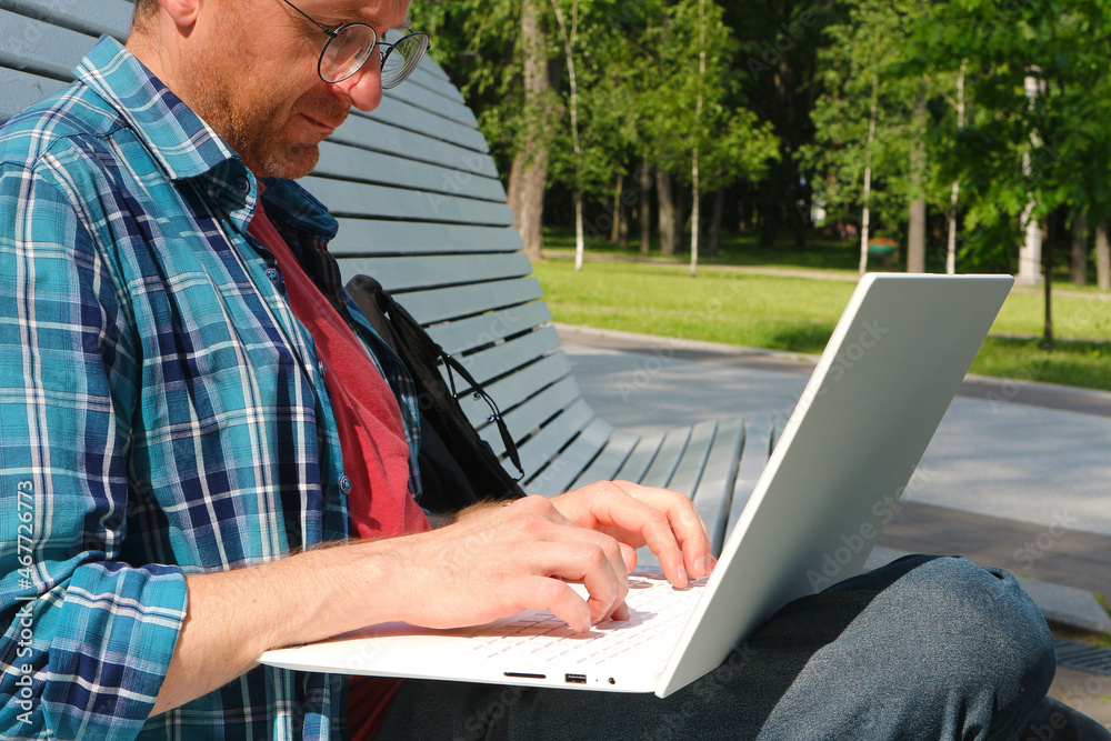 A man in a shirt and glasses is typing on a park bench. A man with a laptop works remotely on the Internet.