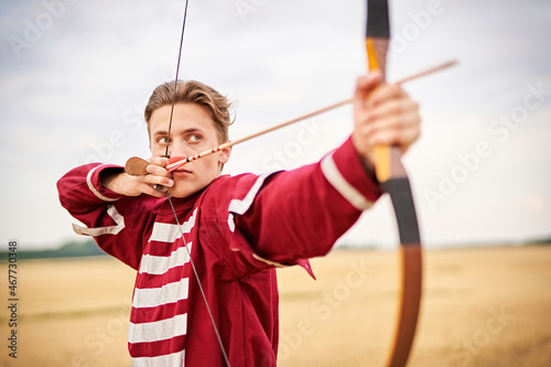 Young male sportsman targeting with bow in a traditional, medieval archer costume - Teenager archer practicing archery in nature - Field archery and recreation concept with a millennial boy