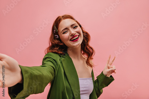 Cute caucasian young girl takes selfie inside pink indoor room. Red-haired lady shows sign of victory by make up lips in red  wearing green business suit. People sincere emotions lifestyle concept.