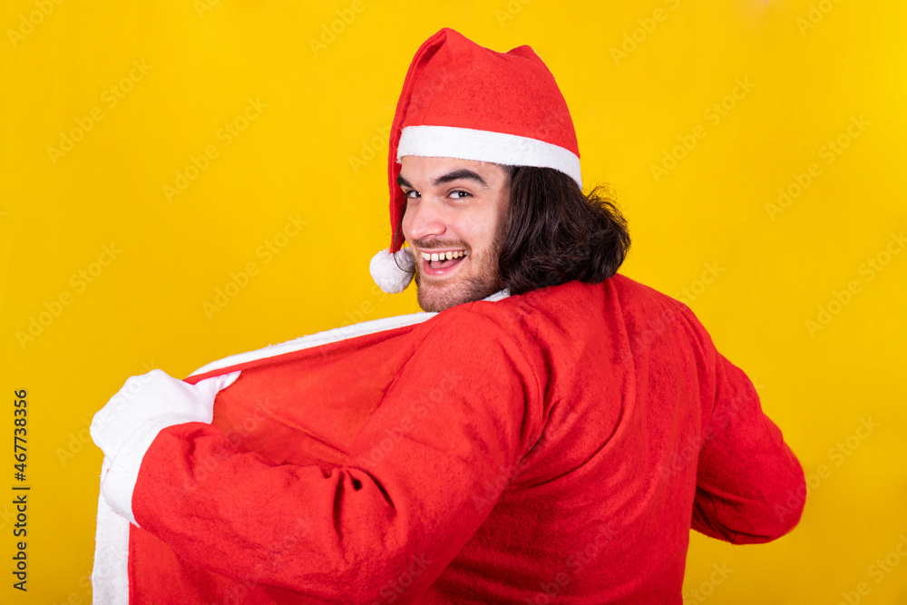 The young man is wearing Santa Claus suit. He smiles and looks towards the viewer with a mischievous look. Yellow background and copy space for your message. The young man is wearing Santa Claus suit. He smiles and looks towards the viewer with a mischievous look. Yellow background and copy space for your message.