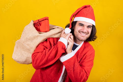 A young man dressed as Santa Claus carries a sack full of presents. He has long hair, a beard and a beautiful smile. Image isolated on yellow background. photo