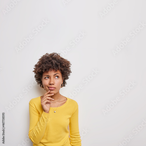 Indoor shot of thoughtful young woman considers something concentrated above with pensive expression makes choice decide how to do dressed casually poses against white background blank space