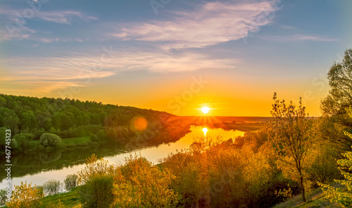 Scenic view at beautiful spring sunset on a shiny river valley with green branches  trees  bushes  grass  golden sun  calm water  deep blue cloudy sky and forest on a background  spring landscape
