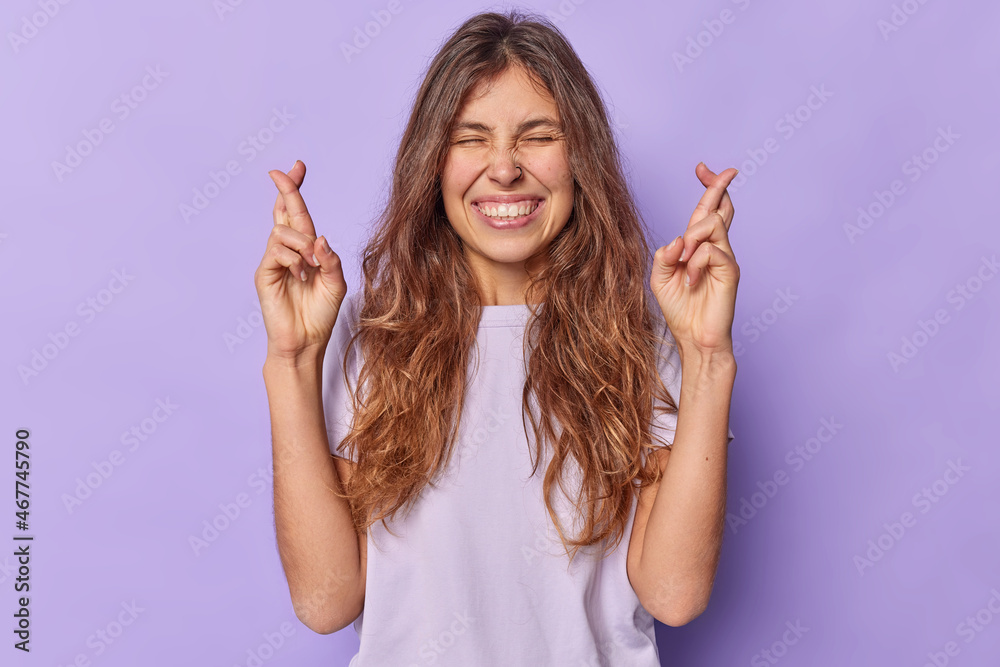 Optimistic young woman anticipates good news keeps fingers crossed begs for positive outcome wears casual t shirt isolated over purple background waits fortune or lucky moment. Body language Optimistic young woman anticipates good news keeps fingers crossed begs for positive outcome wears casual t shirt isolated over purple background waits fortune or lucky moment. Body language