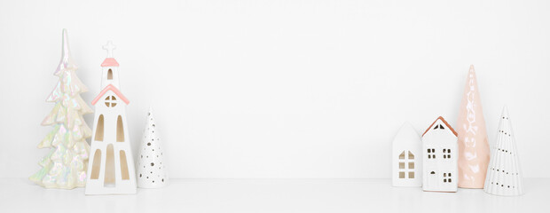 White and soft pink Christmas decor on a white shelf against a white wall banner background with copy space. Ceramic church, houses and shiny trees.
