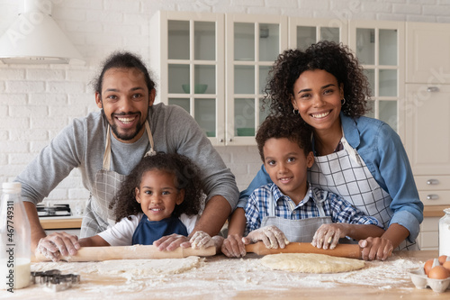 Happy African family baking dessert for dinner together. Sibling children helping couple of parents to cook, rolling dough on table with flour messy, looking at camera, smiling, Head shot portrait