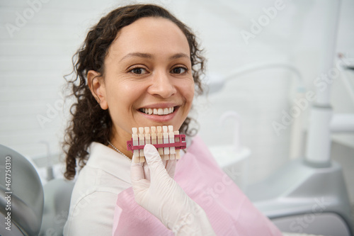 Close-up of dentist hand holding a teeth color chart near the face of a dentistry clinic woman patient smiling with beautiful toothy smile looking at camera. Teeth whitening, bleaching concept