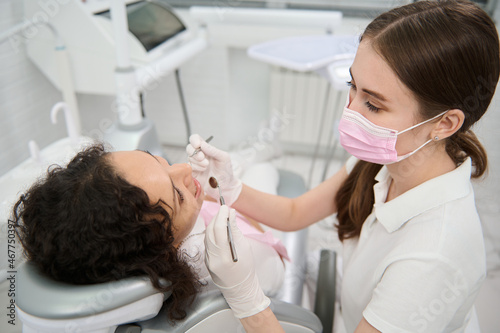 Dentist hygienist in medical protective mask examining the oral cavity of a female patient with sterile stainless steel dental mirror and probe in dentistry clinic during planned medical check-up