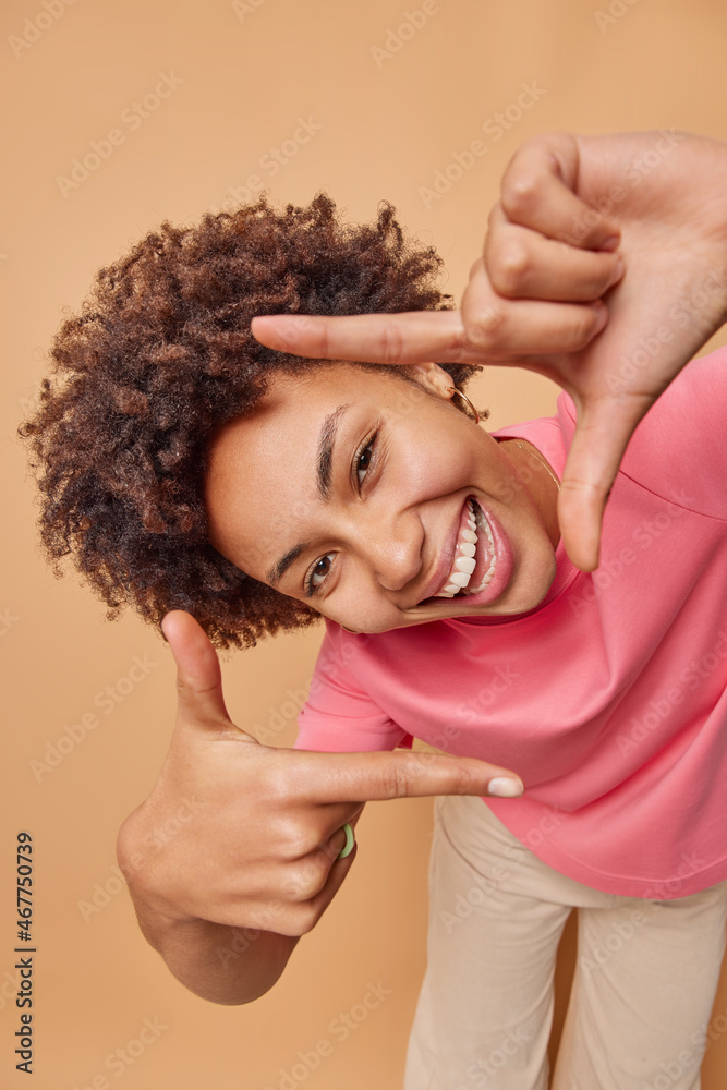Young positive woman makes camera frame with fingers tilts head and smiles happily searches perfect angle has playful mood poses indoor against brown background uses imagination pictures something Young positive woman makes camera frame with fingers tilts head and smiles happily searches perfect angle has playful mood poses indoor against brown background uses imagination pictures something