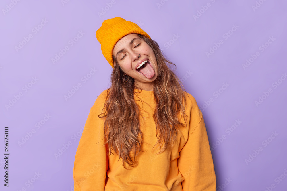 Playful positive young woman tilts head sticks out tongue foolishes around keeps eyes closed wears orange hat and casual jumper isolated over purple background. Human emotions reactions attitude Playful positive young woman tilts head sticks out tongue foolishes around keeps eyes closed wears orange hat and casual jumper isolated over purple background. Human emotions reactions attitude
