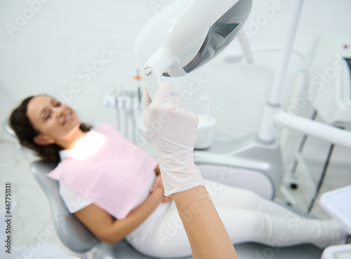 Close-up of a hand in surgical glove adjusting the light against a blurred smiling patient sitting in the dentist's chair and waiting for dental treatment. Medical equipment in dentistry