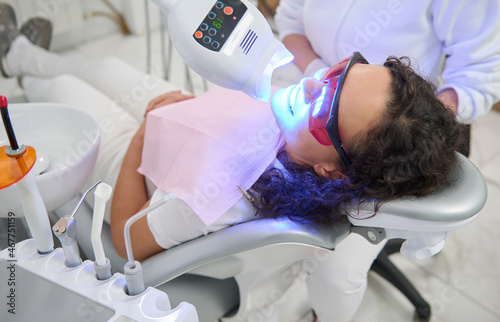 Beautiful young woman patient sitting in dentists chair, with open mouth receiving bleaching treatment with modern special ultraviolet light lamp for teeth whitening procedure in dental clinic