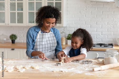 Happy African American mom helping cute daughter to bake biscuit, cut dough for cookies. Young mother and little kid girl cooking bakery food, pastry dessert, snack at table with flour messy on top
