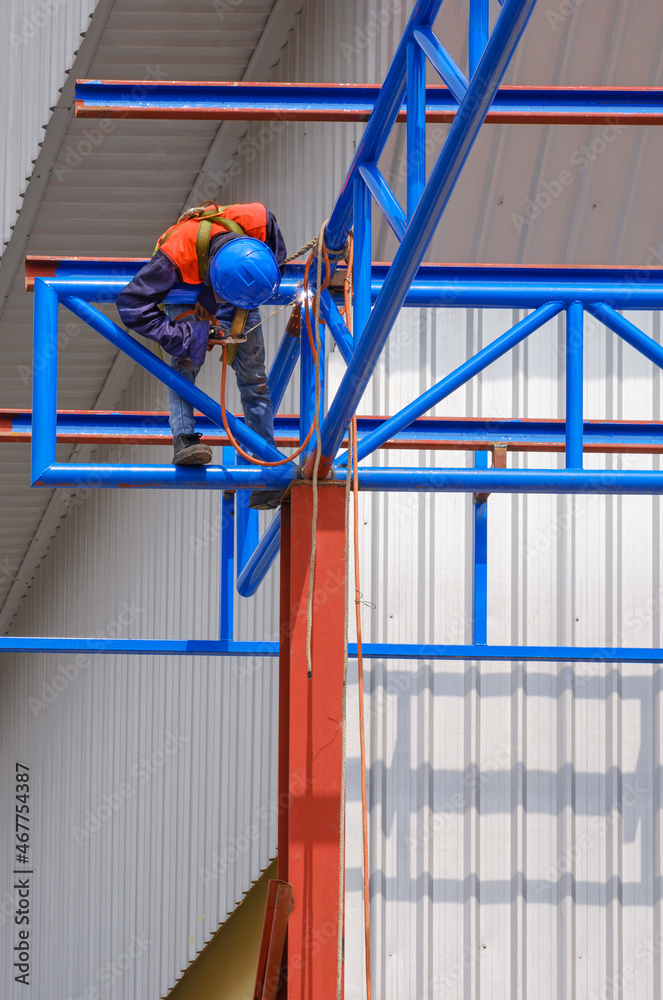 Low angle view of construction worker with safety equipment welding metal roof structure of warehouse building in construction site, work safety concept