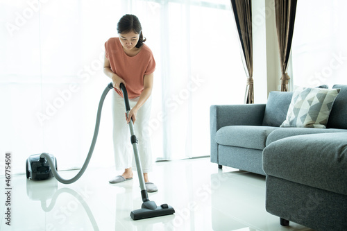 Asian young housekeeper using vacuum machine to clean a dirty floor in the living room.